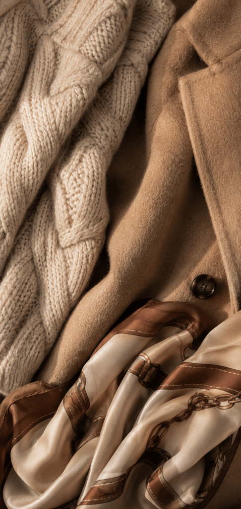 Close-up detail of mixed textures showing a thick cable-knit sweater fabric overlapping with a soft camel wool coat and a silk scarf